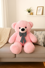 Large pink teddy bear with a black and white checkered bow sitting on a beige sofa with a cozy blanket and cushion in a modern living room