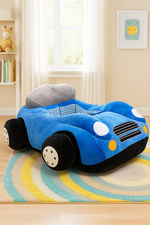 Soft plush kids’ chair in the shape of a blue car with black wheels, white headlights, and a gray headrest, placed on a colorful striped rug in a bright nursery room