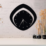 Modern analog wall clock with a sleek black geometric design, mounted on a white brick wall above a minimalist desk setup with a vintage camera and decor items.