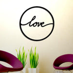 Black MDF circular wall art featuring the word "love" in elegant cursive, mounted on a light-colored wall above green plants and purple modern chairs.