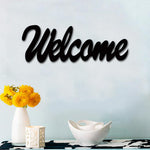 Black MDF wall art featuring the word "Welcome" in elegant cursive typography, mounted on a light blue wall above a table with yellow flowers and decorative bowls.