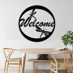 Circular black "Welcome" wall decor with dragonfly accents, mounted on a white wall above a minimalist wooden dining setup.