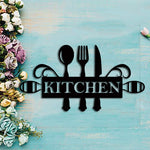 Black cutlery wall art featuring a spoon, fork, and knife design with the word 'KITCHEN' in bold, surrounded by elegant flourishes, displayed on a pastel blue wooden background with floral accents on the side.
