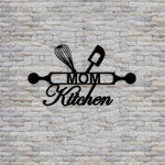 Black MDF wall art featuring the phrase "MOM Kitchen" with a rolling pin, whisk, and spatula design, mounted on a textured stone wall.