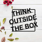 Black MDF wall art with the phrase "THINK OUTSIDE THE BOX" in bold typography, set against a white wooden background with decorative leaves and red flowers.