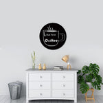 Black round wall art with a coffee cup design and the text 'But first Coffee,' mounted on a light grey wall above a white dresser decorated with books, a lamp, a plant, and small decorative items.
