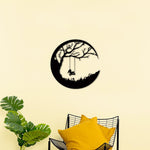 Black swing wall art featuring a circular design with a silhouette of a tree, swing, and child, mounted on a light yellow wall above a chair with yellow cushions.