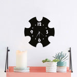 Classic wall clock with a unique black puzzle-piece design, featuring white numerals and sleek hands, mounted on a white wall above a shelf with candles and potted plants