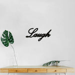 Black MDF wall art featuring the word "Laugh" in elegant cursive typography, mounted on a white wall above a wooden table with minimalist decor and a green leaf in a glass vase.