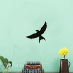 Black bird wall decor featuring a single silhouette of a bird in flight, mounted on a light green wall above a decorative arrangement of books and a vase.