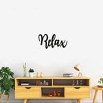 Black MDF wall art with the word "Relax" in elegant cursive typography, mounted on a white wall above a modern wooden console with decorative items and plants