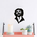 Black wall art featuring a stylized portrait design of a man with a cigar and a beret, mounted on a white wall above a coral shelf adorned with a candle and small potted succulents.