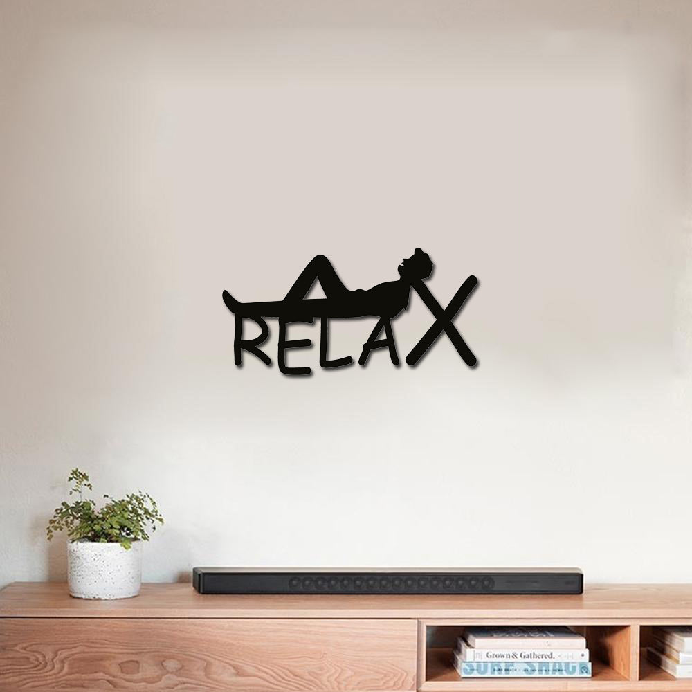 Black MDF wall art with the word "RELAX" featuring a silhouette of a person reclining, mounted on a neutral wall above a wooden console with decor items