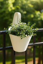 White hanging planter with green leafy plant placed on a black balcony railing with a blurred garden background.