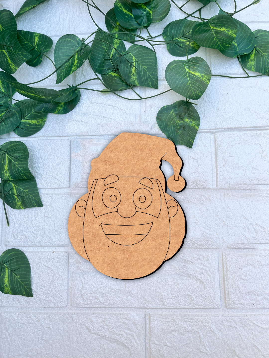 Unpainted DIY MDF Santa face cutout with engraved line art, placed on a white textured brick wall background with green vine leaves.
