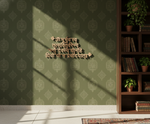 A decorative wall with embossed Sanskrit text in the center, set against olive green patterned wallpaper. Sunlight casts shadows across the wall and floor.