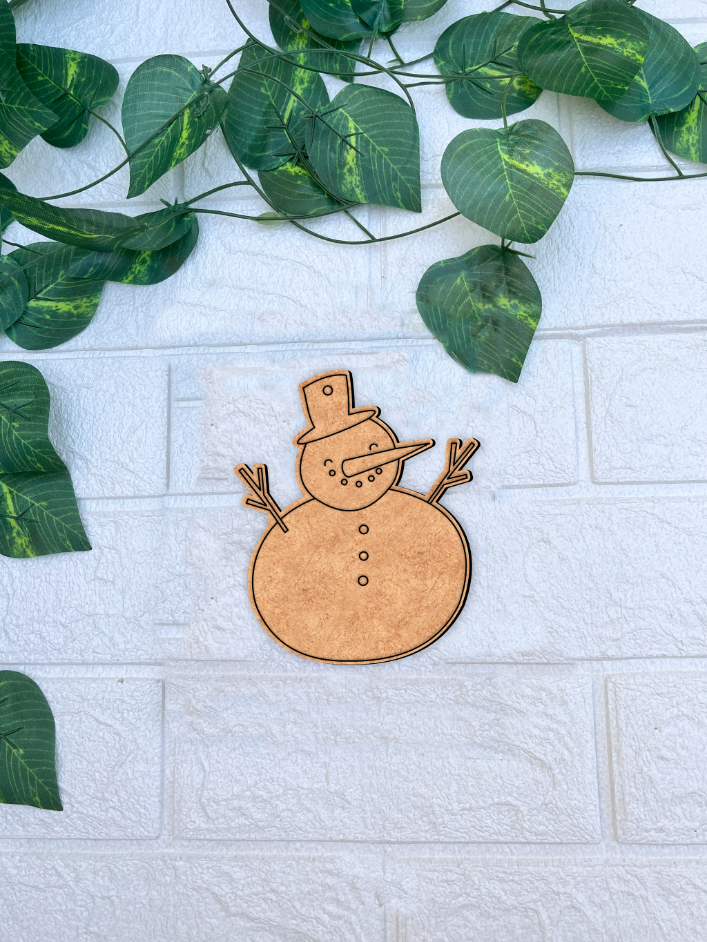 Unpainted MDF snowman DIY cutout with hat, carrot nose and buttons on white brick background with green vine leaves.