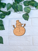 Unpainted MDF snowman DIY cutout with hat, carrot nose and buttons on white brick background with green vine leaves.