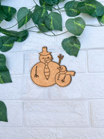 Unpainted MDF snowman and small snowman DIY cutout on white brick background with green vine leaves.