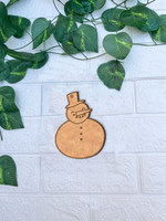 Unpainted MDF snowman DIY cutout with hat, carrot nose and buttons on white brick background with green vine leaves.