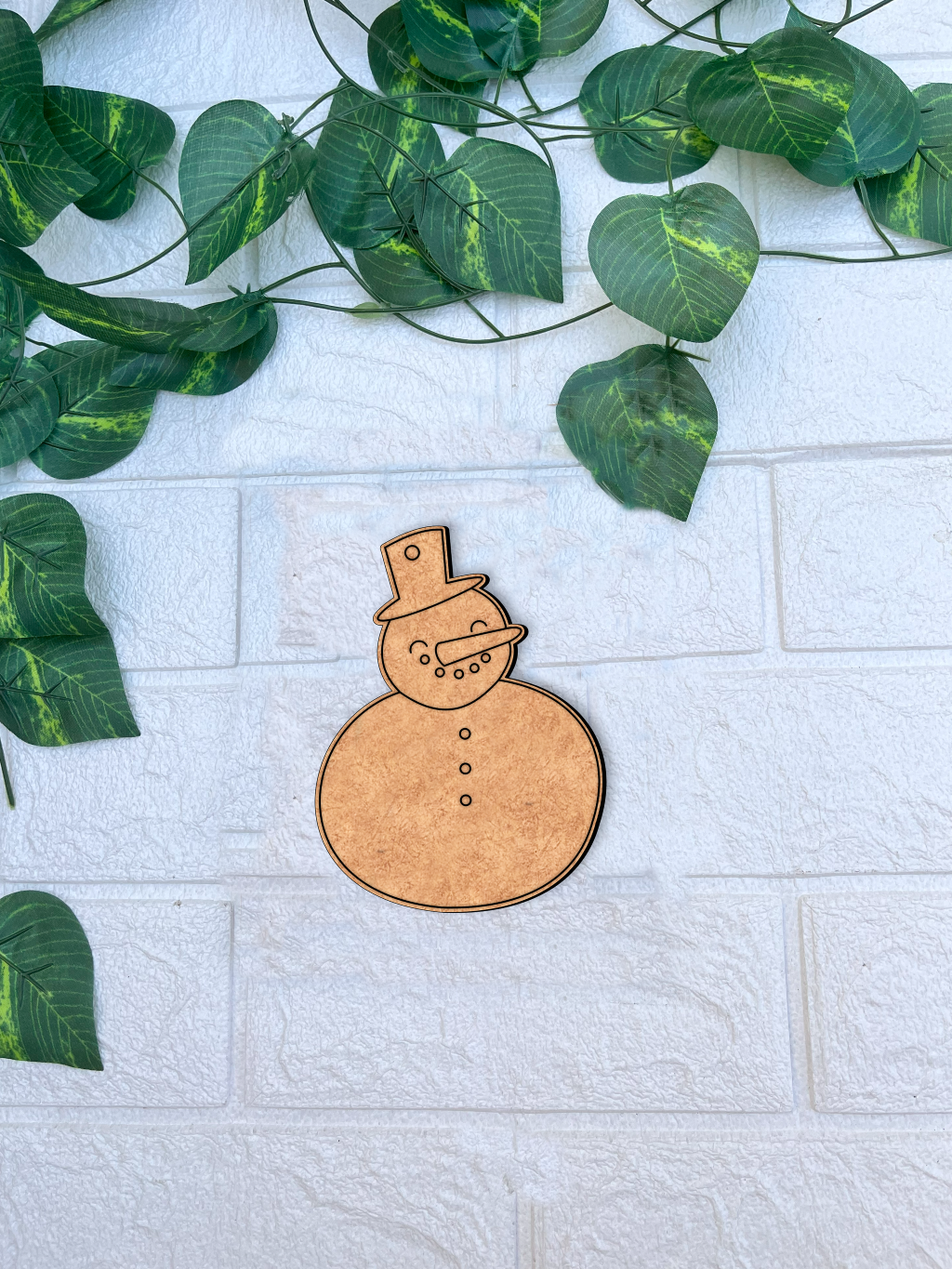 Unpainted MDF snowman DIY cutout with hat, carrot nose and buttons on white brick background with green vine leaves.