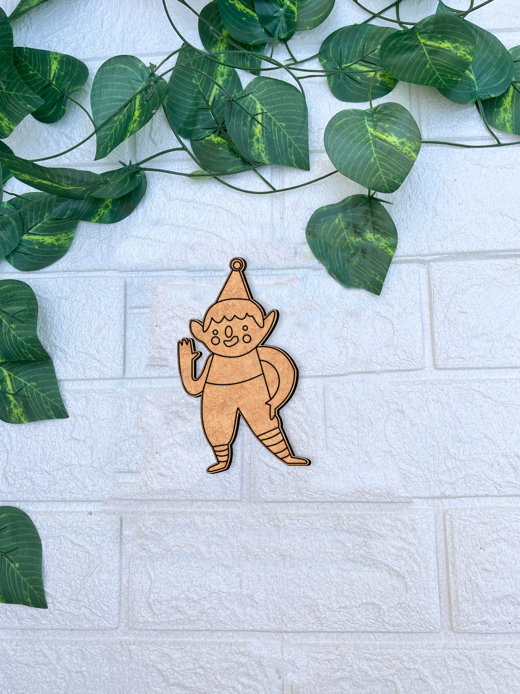Unpainted MDF Christmas elf waving DIY cutout on white brick background with green vine leaves.