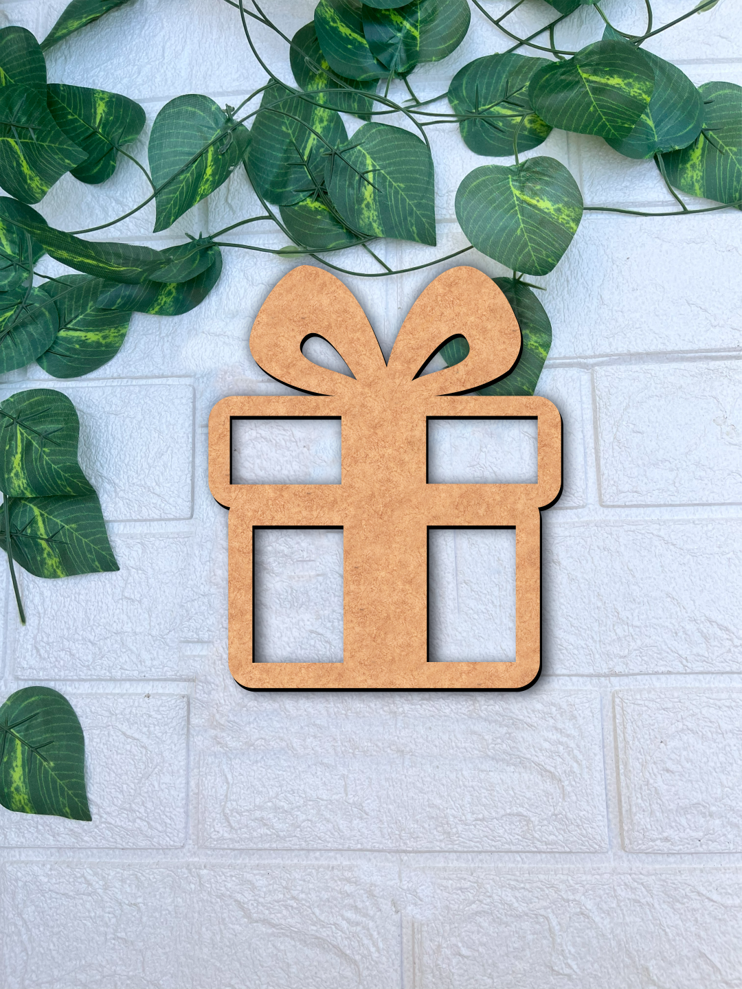 Unpainted DIY MDF gift box cutout with bow and open window design, placed on a white textured brick wall background with green vine leaves.
