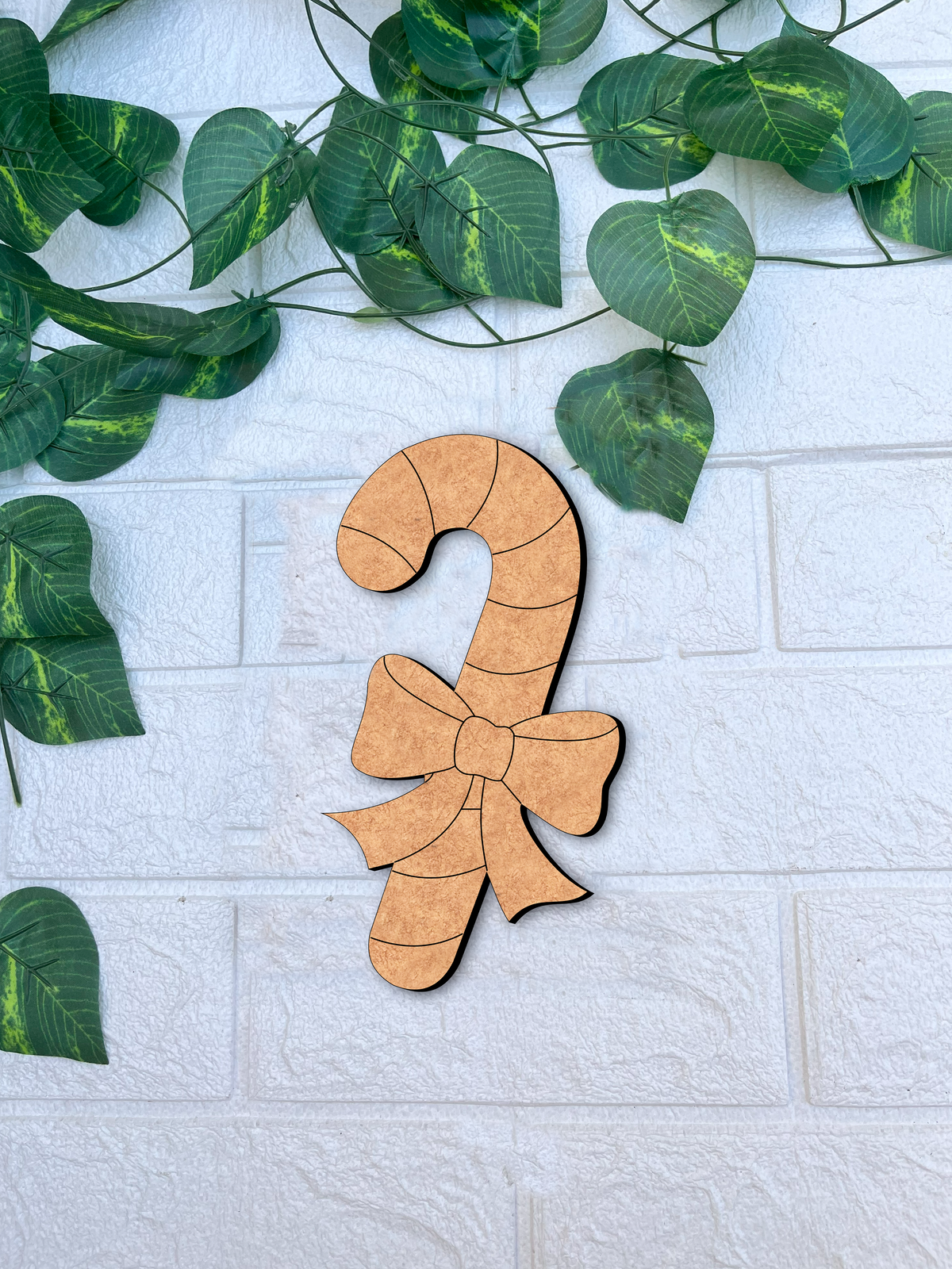 Unpainted DIY MDF candy cane cutout with engraved bow and stripe details, placed on a white textured brick wall background with green vine leaves.