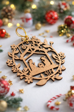 MDF nativity Christmas ornament with Mary, Joseph, and baby Jesus inside a wreath frame, placed on a festive holiday background with decorations and lights.