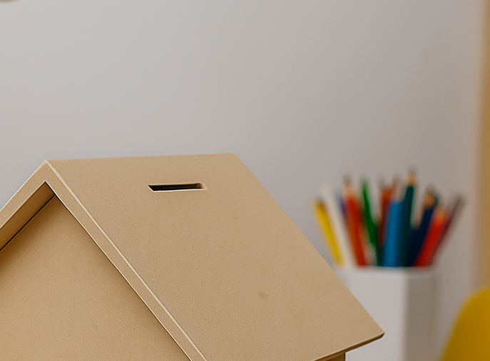 MDF house-shaped piggy bank with coin slot on top, placed indoors near a container of colorful pencils