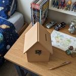 MDF house-shaped piggy bank on a wooden surface with books and toys in the background