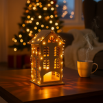 Decorative house-shaped lantern with string lights on a table, with a Christmas tree in the background.
