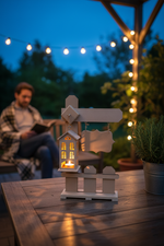 MDF lantern with mini house design on outdoor table, glowing candle, evening lights in background.