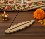 Feather-shaped MDF agarbatti stand with incense smoke, diya and flowers on table.