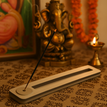 rectangular incense stick holder on patterned cloth, Ganesha idol and diya in background.