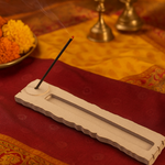 MDF incense holder with burning stick on mandala-print cloth and scattered marigold petals.
