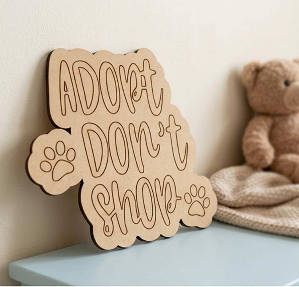MDF DIY sign with 'Adopt don't shop' text next to a teddy bear on a light blue surface.