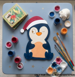 Painted DIY MDF penguin with a Santa hat on a table with art supplies