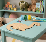 Children's play area with a table featuring a MDF DIY dinosaur craft, paint supplies, and toys on a shelf in the background.