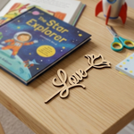 MDF DIY  bookmark 'Love' sign on a wooden surface with a children's book and toy rocket in the background.