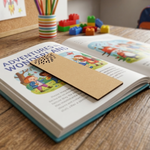 Open book with a MDF DIY  bookmark on a wooden surface, surrounded by colorful toys and stationery.