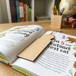 Open book with a MDF DIY bookmark on a wooden table, surrounded by books and stationery items.