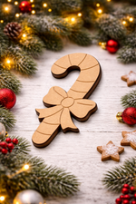 Unpainted DIY MDF candy cane cutout with engraved bow and stripe details, placed on a white wooden surface with festive pine branches, red ornaments, and warm fairy lights.