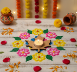 Decorative floral rangoli with a lit candle in a MDF candle holder on a wooden surface, surrounded by colorful pots and flowers.