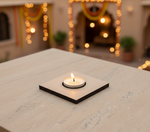 Candle on a square black base with blurred festive lights and decorations in the background with mdf tealight candle holder.
