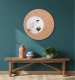 Decorative round mirror with floral patterns on a teal wall above a wooden bench with vases and a plant.