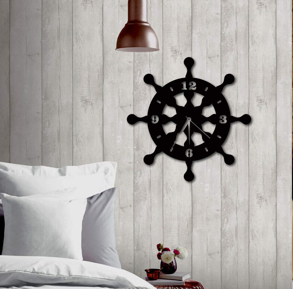 Black MDF wheel-shaped wall clock mounted on a rustic wooden panel wall, styled above a cozy bed with white pillows.