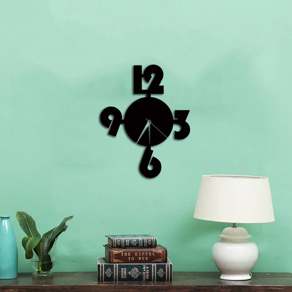 Black modern wall clock with oversized numbers on a light green wall, accompanied by a wooden table with books and a decorative table lamp.