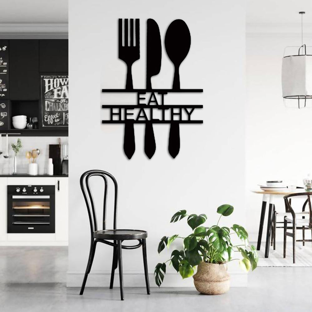 Modern black wall art with the words 'Eat Healthy' designed with cutlery silhouettes, mounted on a bright kitchen wall near a black chair and a green potted plant