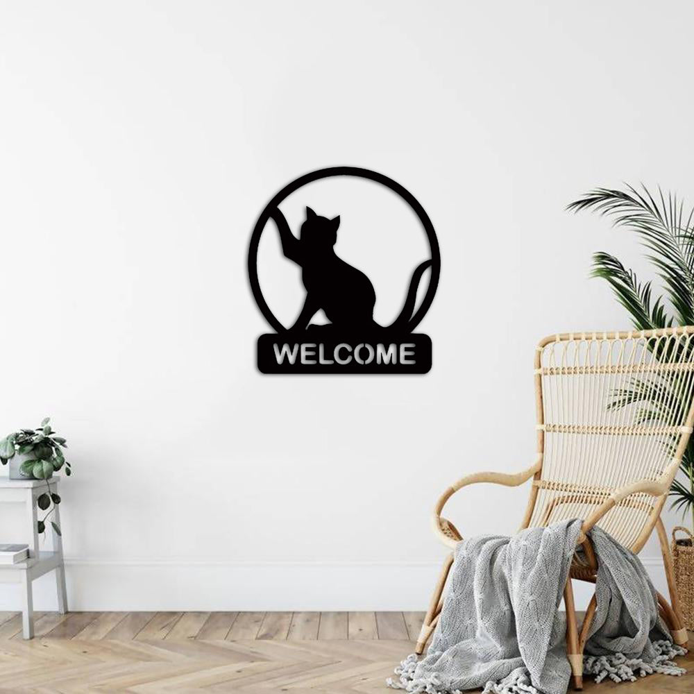 Black cat welcome sign board with a circular frame and the word 'WELCOME,' mounted on a white wall in a cozy living space with a wicker chair, gray throw blanket, and indoor plants.